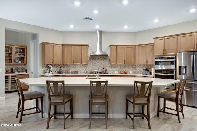 kitchen with appliances with stainless steel finishes, a kitchen bar, wall chimney exhaust hood, tasteful backsplash, and an island with sink