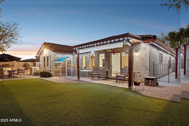 back of house at dusk featuring stucco siding, a patio area, a tiled roof, fence, and a lawn