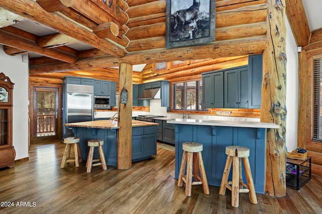 kitchen featuring a breakfast bar area, log walls, dark hardwood / wood-style flooring, built in appliances, and wall chimney exhaust hood