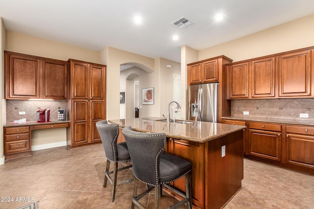 kitchen with a breakfast bar, an island with sink, stainless steel refrigerator with ice dispenser, brown cabinetry, and a sink