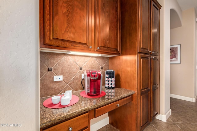 kitchen with tile patterned flooring, backsplash, baseboards, brown cabinets, and dark stone countertops