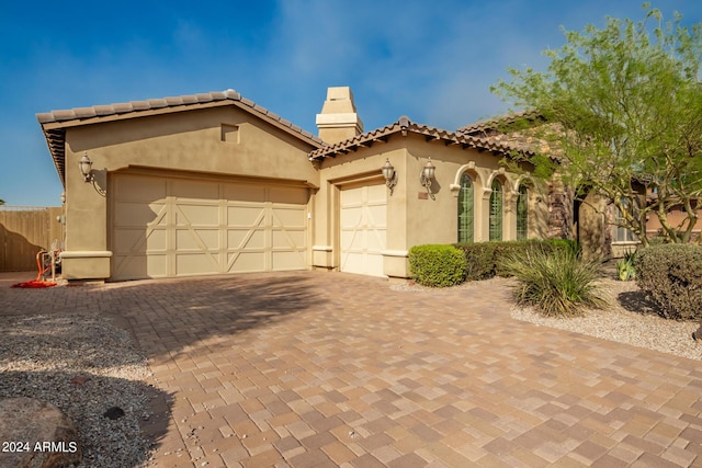 mediterranean / spanish house featuring a tile roof, stucco siding, a chimney, decorative driveway, and an attached garage