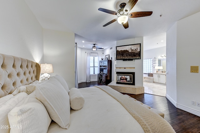 bedroom with dark wood-style flooring, baseboards, a multi sided fireplace, ensuite bath, and a ceiling fan