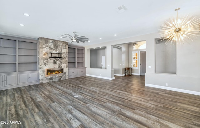 unfurnished living room featuring ceiling fan with notable chandelier, dark wood-type flooring, built in shelves, and a stone fireplace