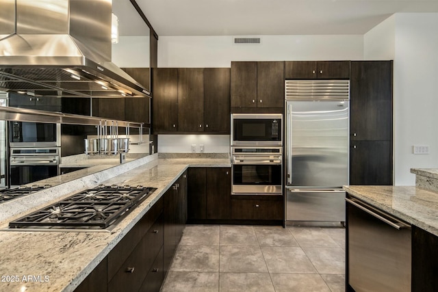 kitchen with light stone countertops, wall chimney exhaust hood, dark brown cabinets, light tile patterned floors, and built in appliances