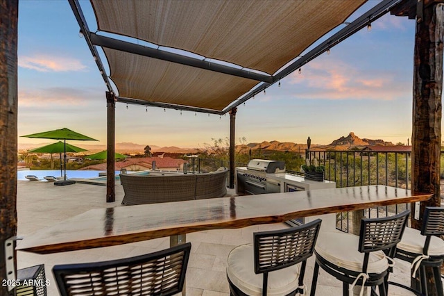 patio terrace at dusk with exterior kitchen, outdoor dining area, a mountain view, and area for grilling