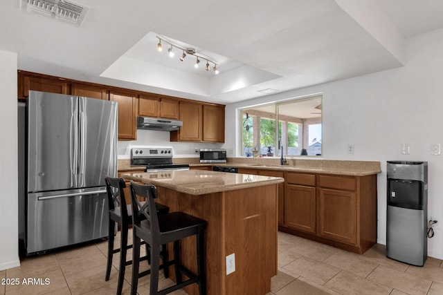 kitchen featuring appliances with stainless steel finishes, under cabinet range hood, a raised ceiling, light tile patterned flooring, and brown cabinets