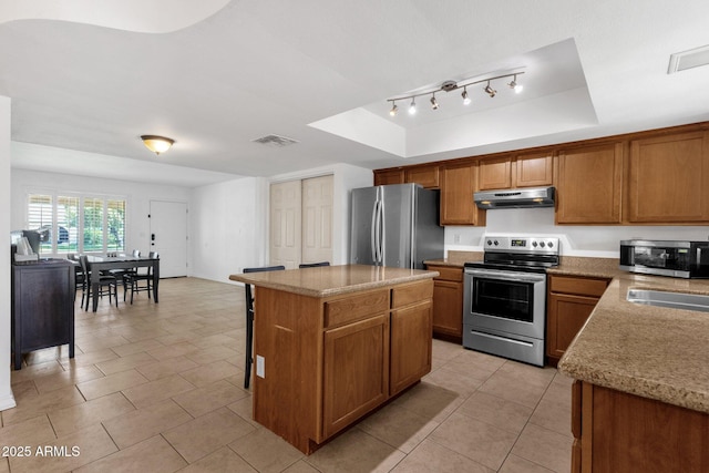 kitchen featuring stainless steel appliances, under cabinet range hood, a raised ceiling, brown cabinets, and a center island