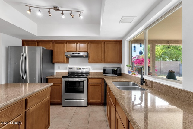 kitchen featuring appliances with stainless steel finishes, a sink, under cabinet range hood, a tray ceiling, and brown cabinets