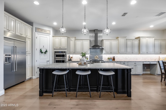 kitchen with light stone countertops, wall chimney range hood, light hardwood / wood-style floors, a kitchen island with sink, and appliances with stainless steel finishes