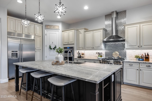 kitchen featuring light hardwood / wood-style floors, wall chimney range hood, stainless steel appliances, and a kitchen island with sink