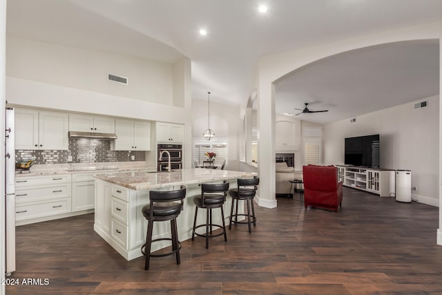 kitchen with a kitchen island with sink, ceiling fan, decorative light fixtures, dark hardwood / wood-style flooring, and stainless steel double oven