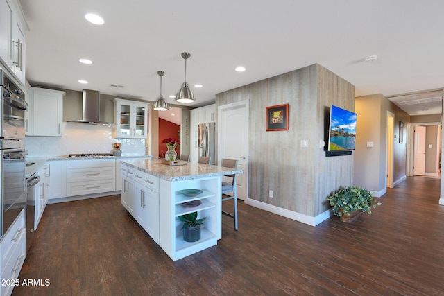 kitchen featuring a center island, hanging light fixtures, wall chimney range hood, white cabinets, and appliances with stainless steel finishes