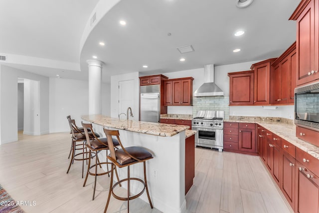 kitchen with a breakfast bar, an island with sink, built in appliances, light stone counters, and wall chimney exhaust hood