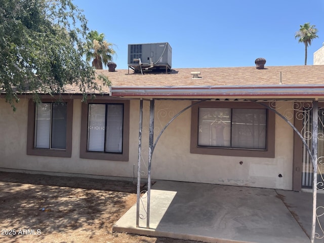 rear view of property with a shingled roof, stucco siding, a patio, and central air condition unit