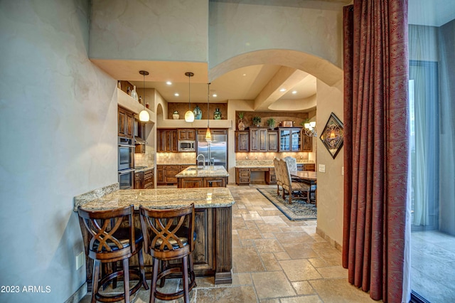 kitchen featuring tasteful backsplash, built in appliances, a kitchen island with sink, light stone countertops, and light tile flooring