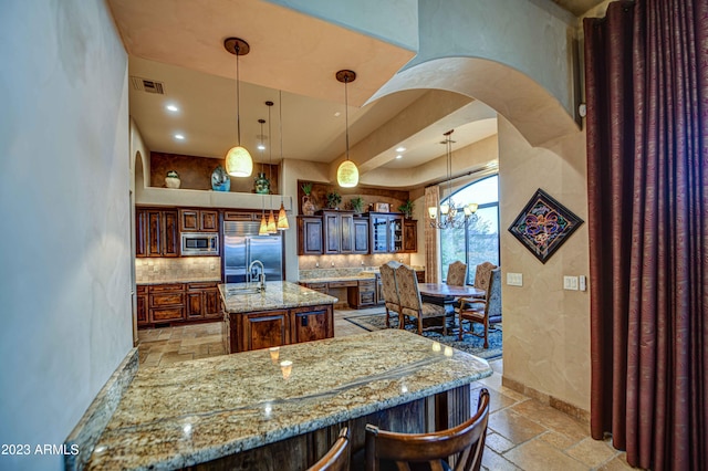 kitchen with an inviting chandelier, built in appliances, an island with sink, light stone countertops, and hanging light fixtures