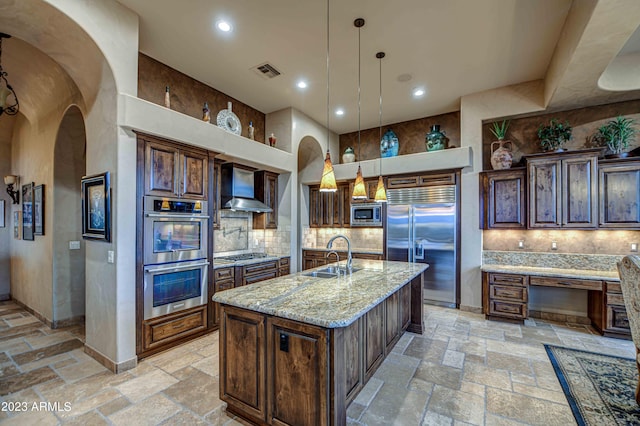 kitchen featuring an island with sink, built in appliances, wall chimney exhaust hood, backsplash, and sink