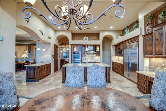 kitchen featuring pendant lighting, built in appliances, a center island with sink, wall chimney exhaust hood, and backsplash