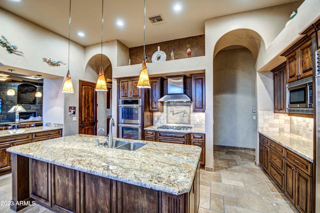 kitchen with hanging light fixtures, appliances with stainless steel finishes, an island with sink, wall chimney range hood, and tasteful backsplash