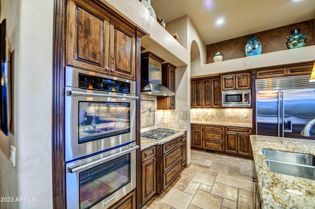 kitchen with built in appliances, sink, light stone counters, backsplash, and wall chimney range hood