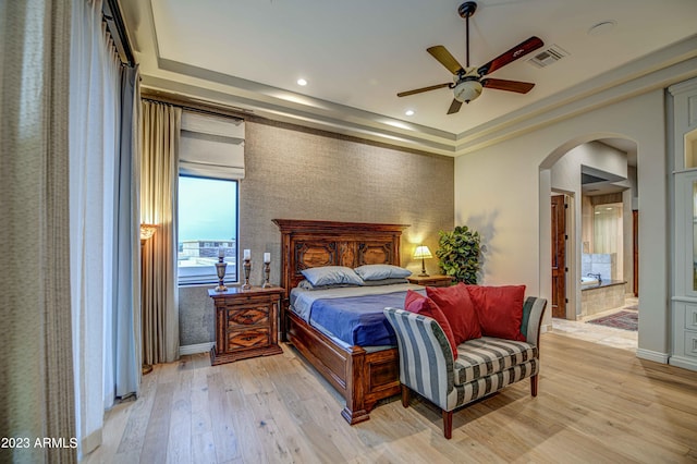 bedroom featuring light hardwood / wood-style flooring, ceiling fan, ensuite bath, and a tray ceiling
