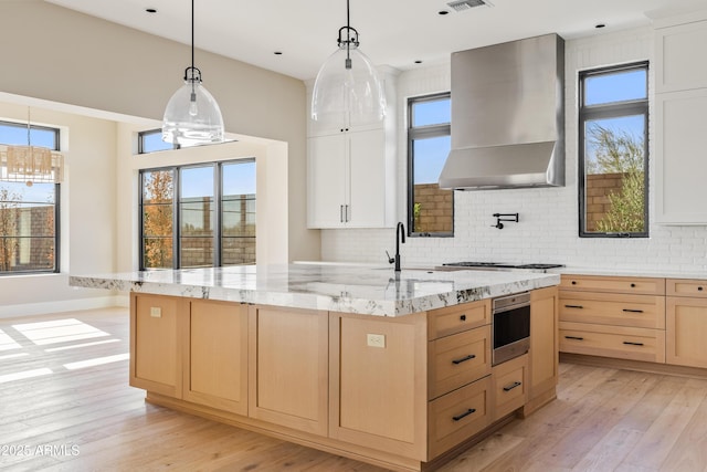 kitchen featuring white cabinets, an island with sink, decorative light fixtures, and wall chimney range hood