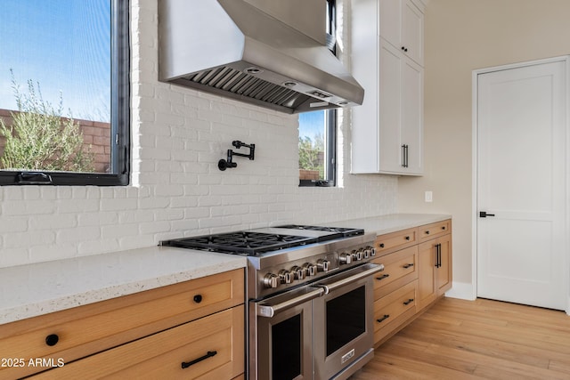 kitchen featuring range with two ovens, wall chimney range hood, light stone countertops, light hardwood / wood-style floors, and white cabinetry