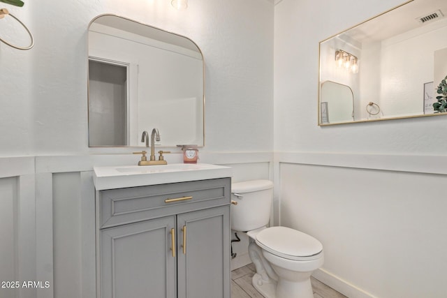 half bathroom featuring toilet, a decorative wall, a wainscoted wall, vanity, and visible vents