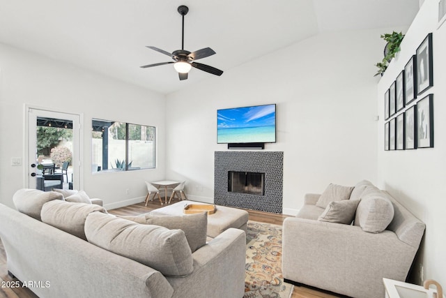 living area with lofted ceiling, a tiled fireplace, ceiling fan, wood finished floors, and baseboards