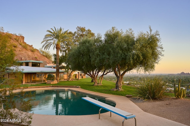 pool at dusk featuring a lawn and a diving board