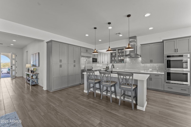 kitchen with pendant lighting, wall chimney range hood, a kitchen breakfast bar, stainless steel appliances, and an island with sink