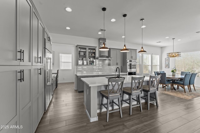 kitchen with wall chimney exhaust hood, gray cabinetry, a center island with sink, pendant lighting, and backsplash