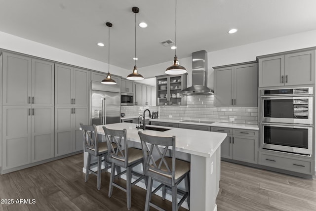 kitchen featuring sink, built in appliances, an island with sink, pendant lighting, and wall chimney range hood