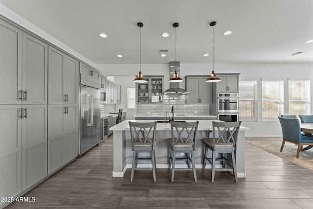 kitchen featuring hanging light fixtures, an island with sink, appliances with stainless steel finishes, and wall chimney range hood