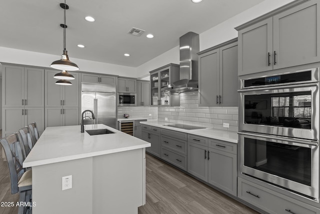 kitchen featuring a kitchen bar, wall chimney exhaust hood, built in appliances, decorative light fixtures, and a kitchen island with sink