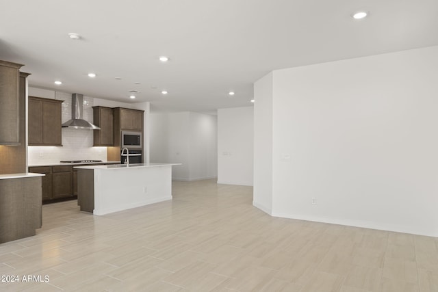 kitchen featuring decorative backsplash, a kitchen island with sink, stainless steel microwave, wall chimney exhaust hood, and light hardwood / wood-style flooring