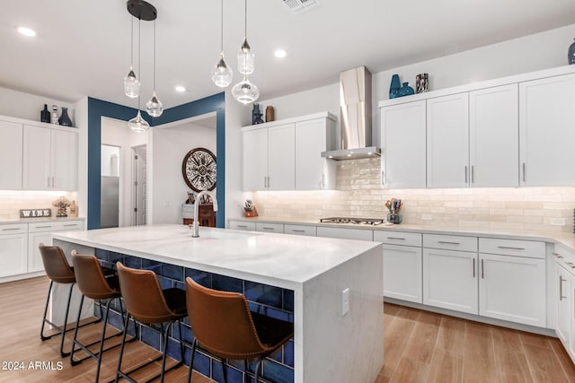 kitchen featuring stainless steel gas stovetop, white cabinetry, a kitchen island with sink, and wall chimney range hood