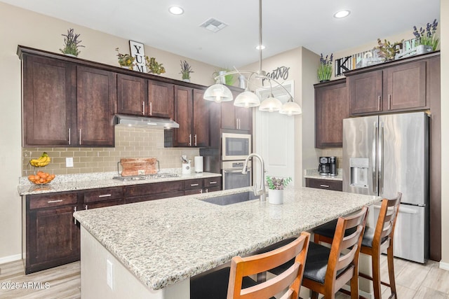 kitchen with pendant lighting, stainless steel appliances, light stone counters, and a center island with sink