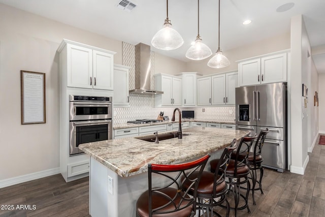 kitchen featuring appliances with stainless steel finishes, pendant lighting, wall chimney range hood, a kitchen island with sink, and white cabinets