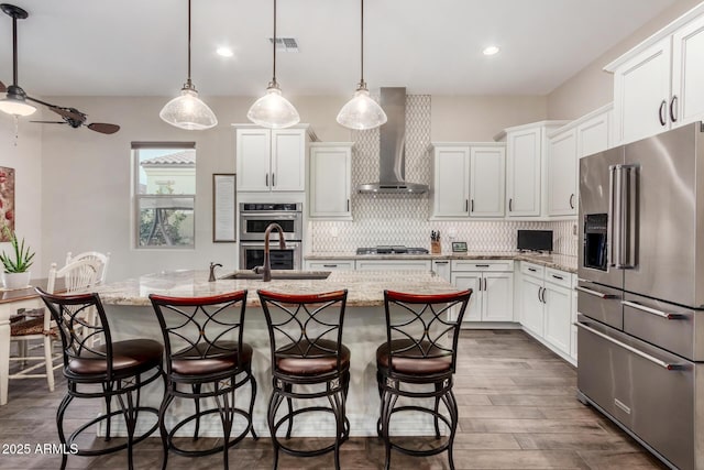 kitchen with wall chimney exhaust hood, sink, appliances with stainless steel finishes, pendant lighting, and a kitchen island with sink