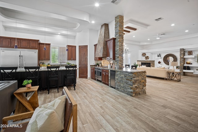 kitchen with a towering ceiling, custom exhaust hood, stainless steel appliances, light hardwood / wood-style flooring, and a fireplace