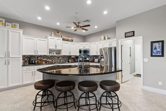 kitchen featuring appliances with stainless steel finishes, light tile patterned floors, a center island with sink, dark stone countertops, and white cabinetry