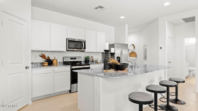 kitchen with light tile patterned floors, a kitchen island with sink, stainless steel appliances, light stone countertops, and white cabinets