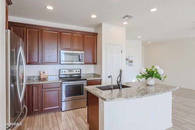 kitchen featuring a kitchen island with sink, light stone counters, stainless steel appliances, sink, and light hardwood / wood-style flooring