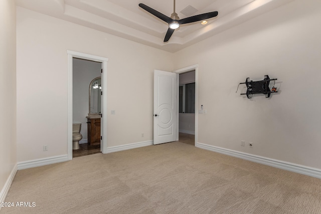 unfurnished bedroom featuring a raised ceiling, light carpet, ceiling fan, and ensuite bathroom