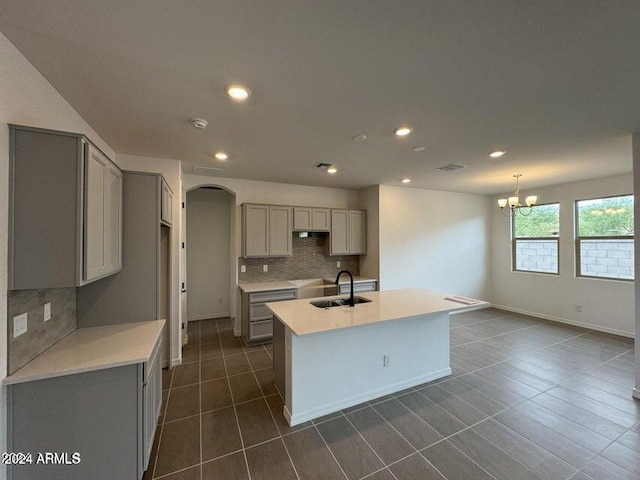 kitchen featuring gray cabinetry, sink, decorative backsplash, an island with sink, and a chandelier