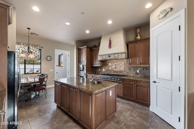 kitchen with sink, appliances with stainless steel finishes, light stone counters, custom range hood, and an island with sink