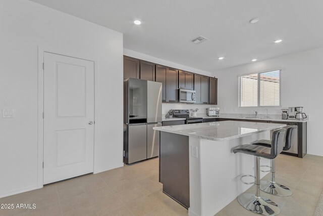 kitchen featuring dark brown cabinetry, light stone countertops, a breakfast bar, a kitchen island, and appliances with stainless steel finishes