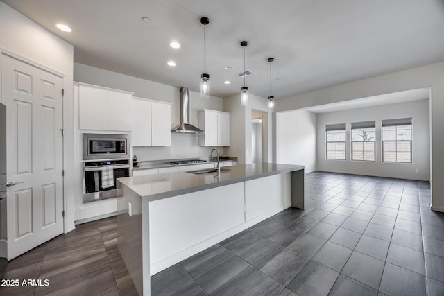 kitchen with wall chimney range hood, an island with sink, appliances with stainless steel finishes, white cabinets, and a sink